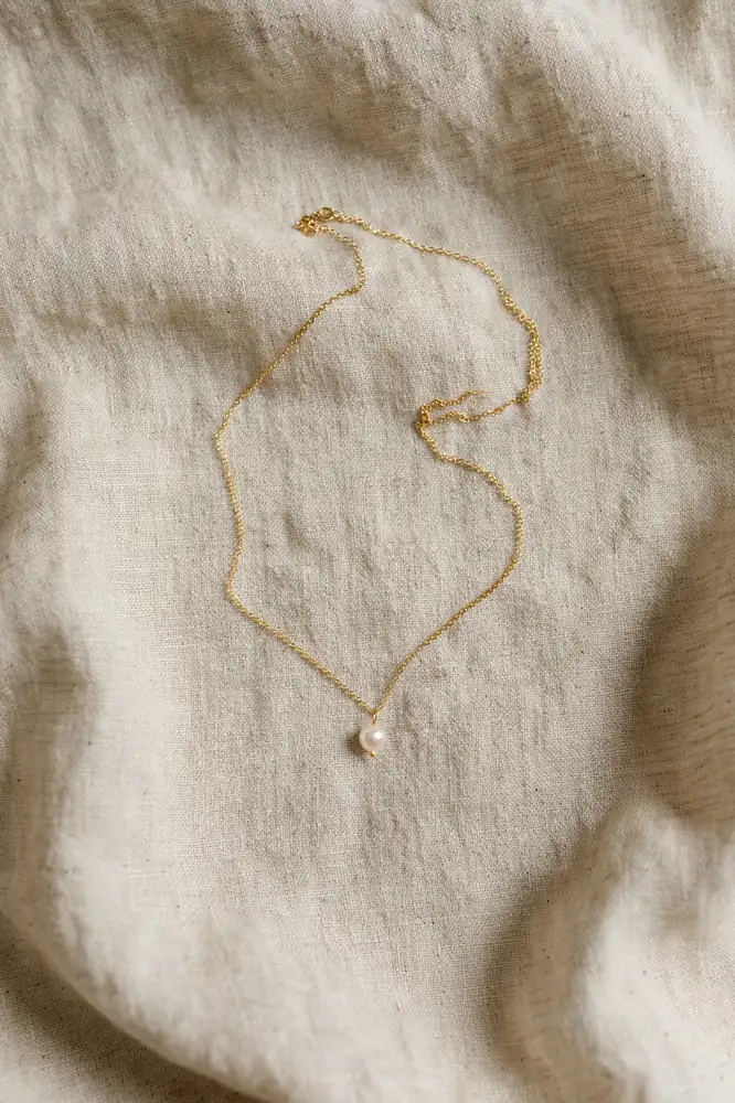 Dainty gold pendant necklace with a tiny pearl on beige linen